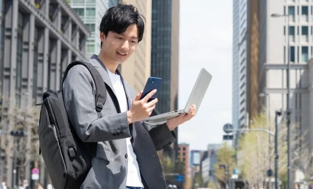 街中でパソコンと携帯を持って立っている男性