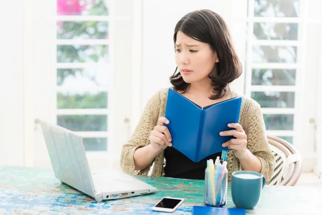 本を読みながらパソコンの前で困惑している女性