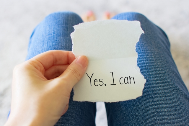 Yes I can と書かれた紙を持って座っている女性