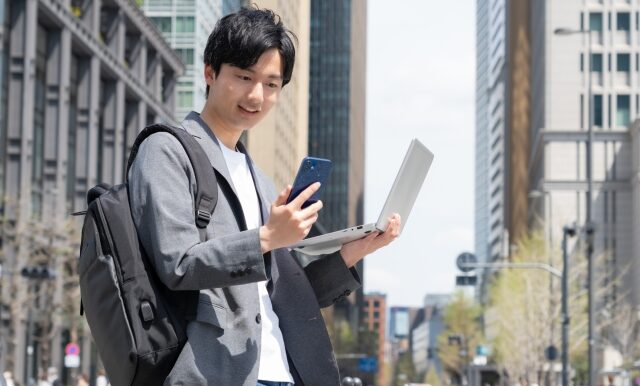 街中でパソコンと携帯を持って立っている男性