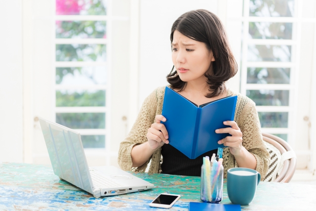 本を読みながらパソコンの前で困惑している女性